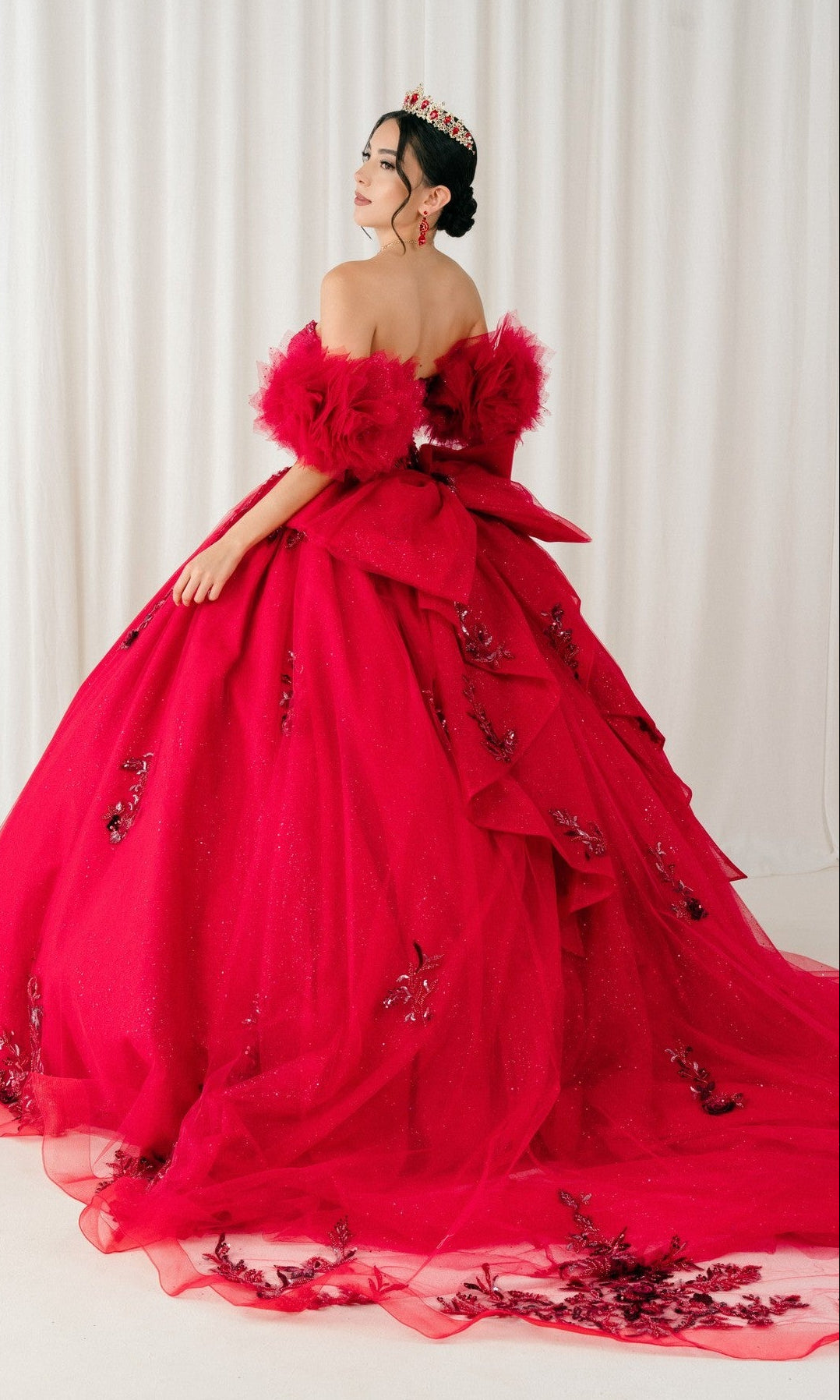 A woman stands before white curtains in the Quinceanera Dress 8380J By Cinderella Couture—an off-the-shoulder bright red ball gown with a corset bodice, floral embellishments, large back bow, and a matching red tiara.
